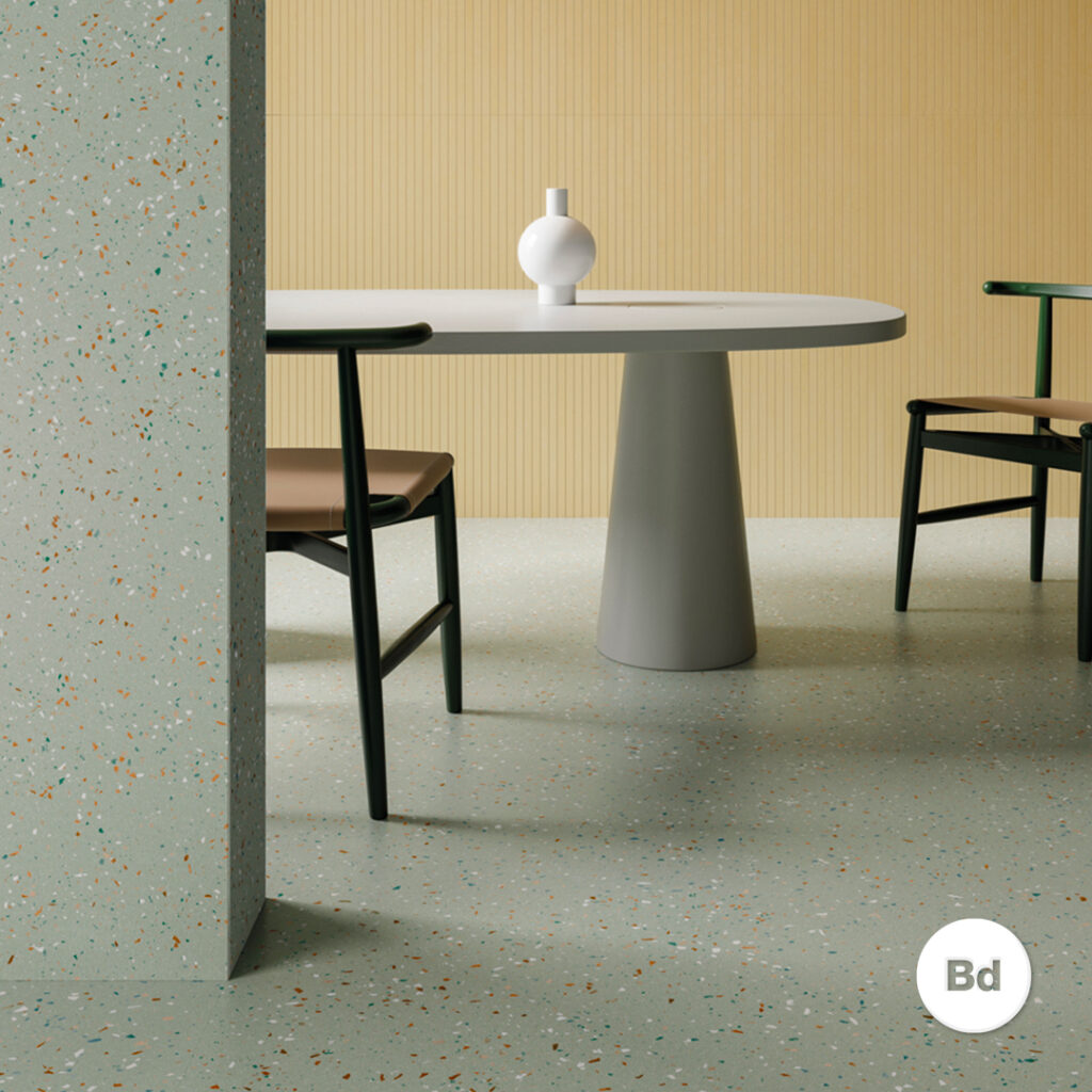 Round white table with a conical gray pedestal base in a minimalist dining nook; beige chairs and terrazzo floor with a yellow panel wall. The table holds a small white vase.
